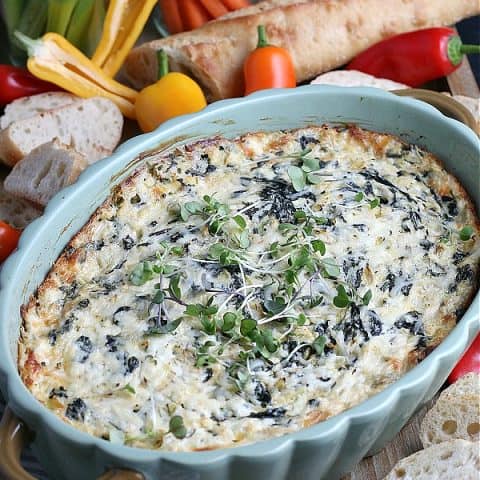 spinach artichoke dip in green baking dish on board with vegetables and bread