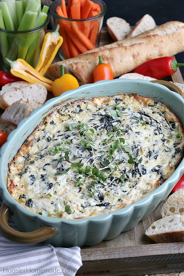 spinach artichoke dip in green baking dish on board with vegetables and bread 