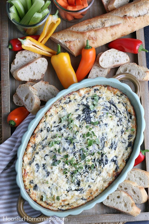 spinach artichoke dip in green baking dish on board with vegetables and bread 