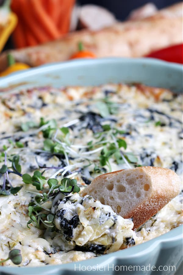 spinach artichoke dip in green baking dish on board with vegetables and bread 