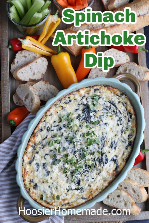 spinach artichoke dip in green baking dish on board with vegetables and bread 